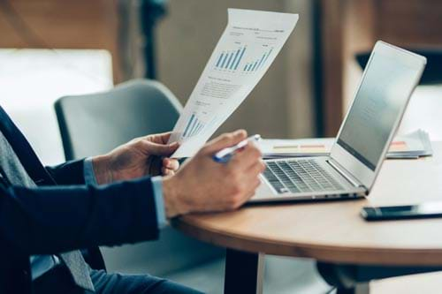 person holding paper with charts sitting by a computer